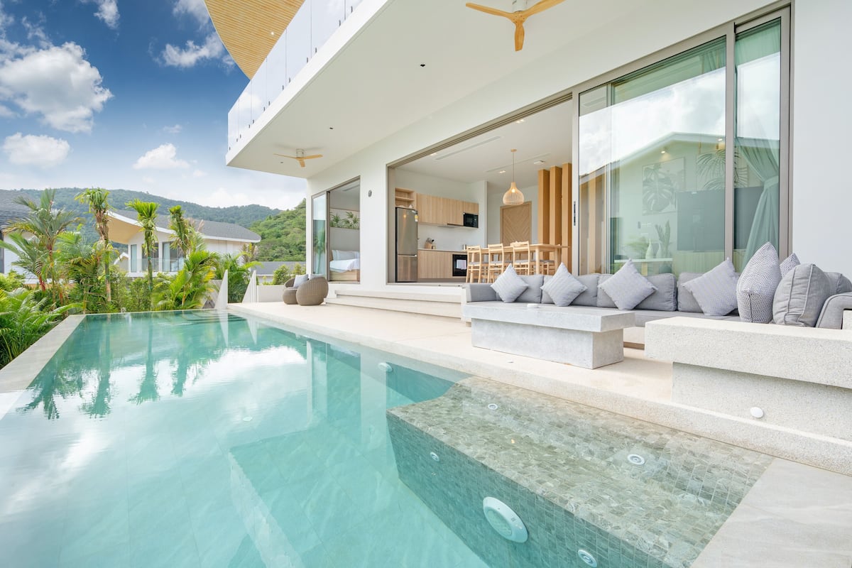 Une maison moderne dotée d’une piscine, d’un espace salon extérieur et d’une cuisine visible à travers les portes vitrées.