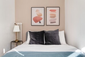 Cama bem arrumada com roupa de cama branca e azul, um travesseiro cinza escuro e um travesseiro branco. Há uma luminária com cúpula branca sobre uma mesa de cabeceira. Duas pinturas abstratas pendem na parede acima da cama.