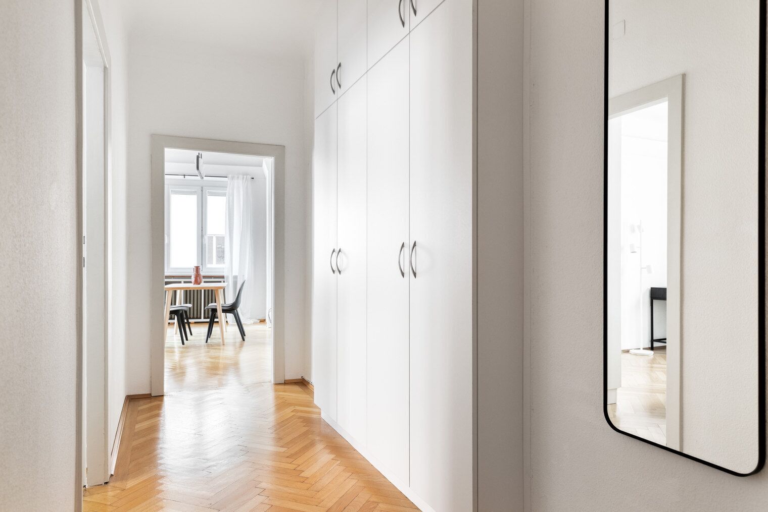 A hallway with a wooden floor, white walls, and a large mirror on the right. There is a door leading to a room with a table and chairs.