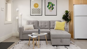 Un salon moderne avec un canapé d’angle gris, une petite table blanche et un réfrigérateur blanc. Deux œuvres d’art encadrées ornent le mur, ainsi qu’une lampe sur pied et une plante en pot.