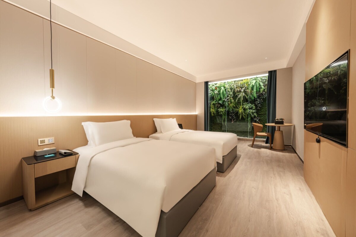 Ein modernes Hotelzimmer mit zwei Betten, einem Nachttisch, einem Schreibtisch, einem Sessel, einem Fernseher und einem großen Fenster mit Blick auf Grünflächen.