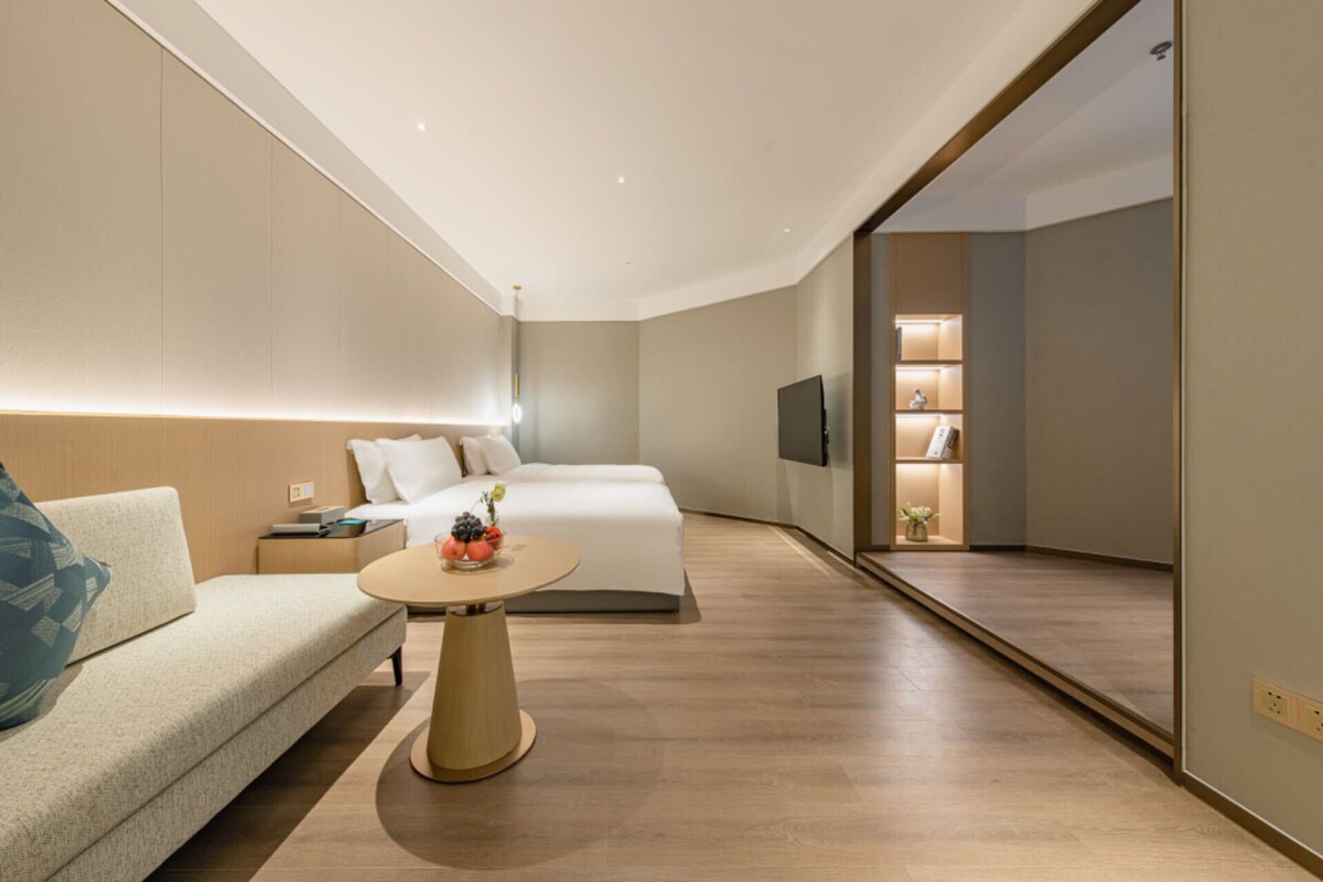 Ein modernes Hotelzimmer mit einer Couch, einem Bett, einem Nachttisch mit Lampe und einem kleinen runden Tisch mit Obst.