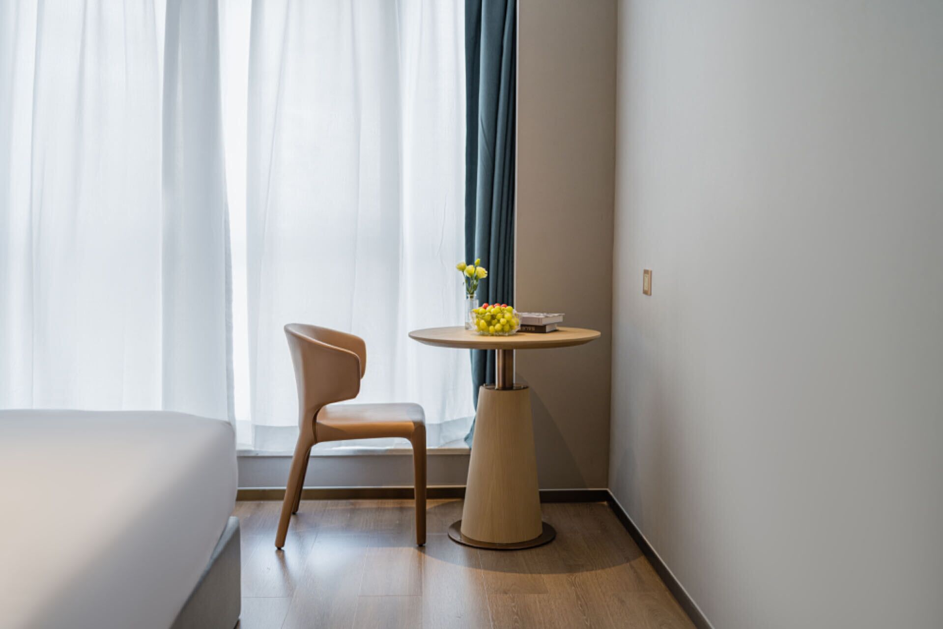 A hotel room with a bed, a chair, a round table with a vase of yellow flowers, and a window with curtains.