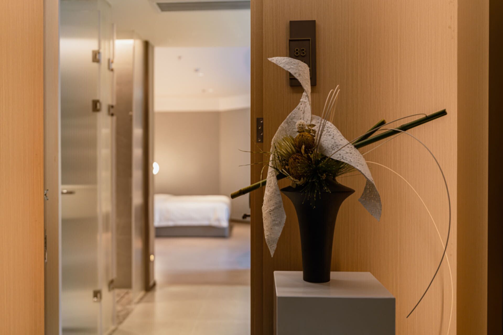 A hotel hallway with a reception desk, a bed in the background, and a decorative plant on a pedestal.
