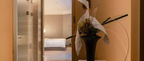 A hotel hallway with a reception desk, a bed in the background, and a decorative plant on a pedestal.