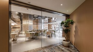 Ein modernes Interieur mit Glastrennwand, einem Essbereich mit Tischen und Stühlen sowie einer Topfpflanze.