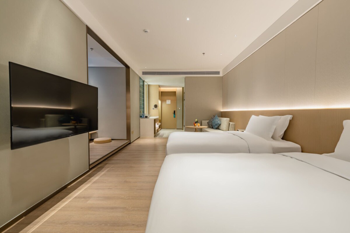 Ein modernes Hotelzimmer mit zwei Betten, einem Flachbildfernseher, einem kleinen Tisch und einem durchgehenden Türspalt einsehbarer Badezimmer.