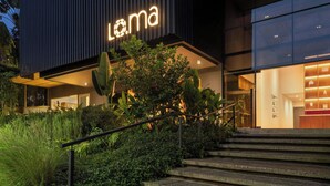 En modern byggnad med en framträdande skylt som visar ordet ”Loma”, en trappa som leder upp till entrén och frodig grönska runt byggnadens bas.