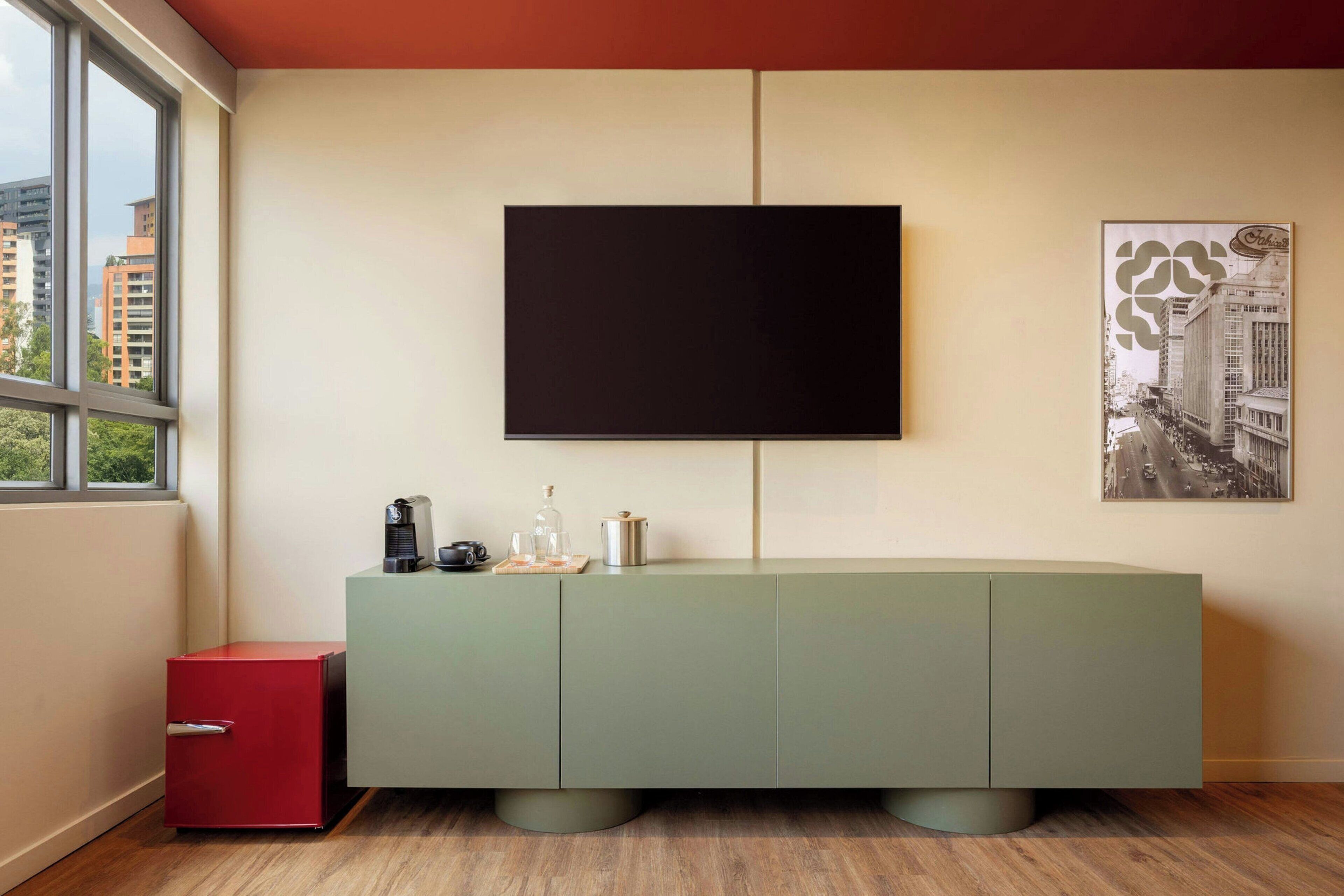 A modern living room with a large flat-screen TV mounted on the wall, a green cabinet with drawers, a red side cabinet, and a framed cityscape picture.