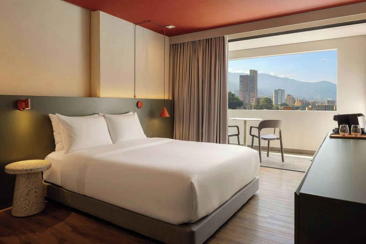 Habitación de hotel con cama, una mesita redonda, una silla, un escritorio y vistas a un paisaje urbano a través de un ventanal grande.