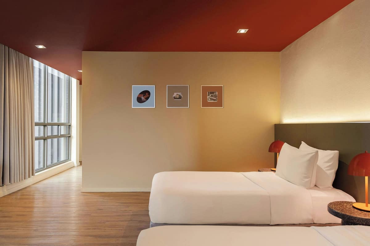 Una habitación de hotel con una cama, dos mesitas de noche, un ventanal con cortinas y tres cuadros enmarcados en la pared.
