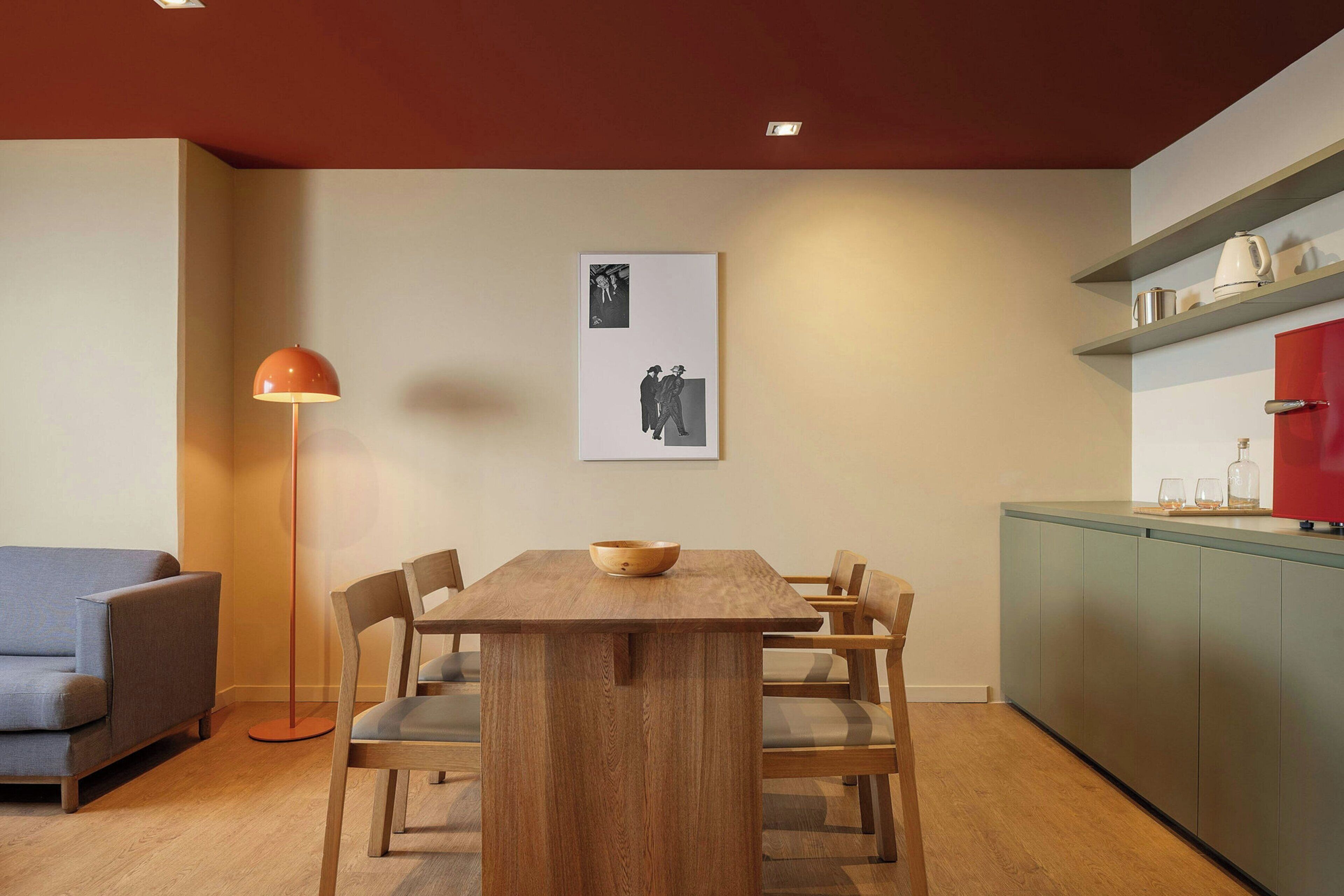Uma área de refeições com uma mesa de madeira, cadeiras, um abajur, um quadro emoldurado e uma cozinha com um armário vermelho.