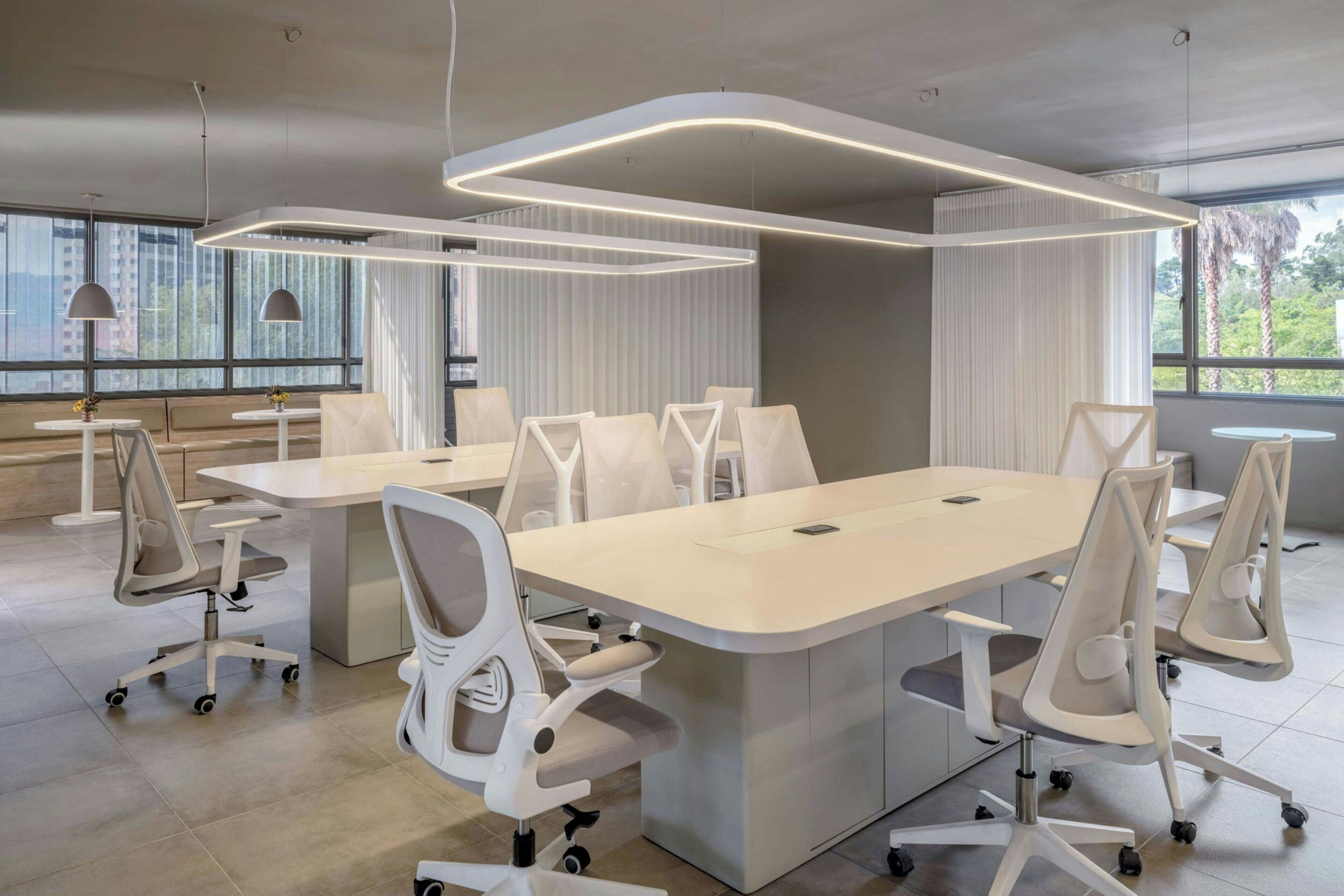 Una sala de conferencias moderna con mesas y sillas blancas, ventanas amplias y vistas a zonas verdes.