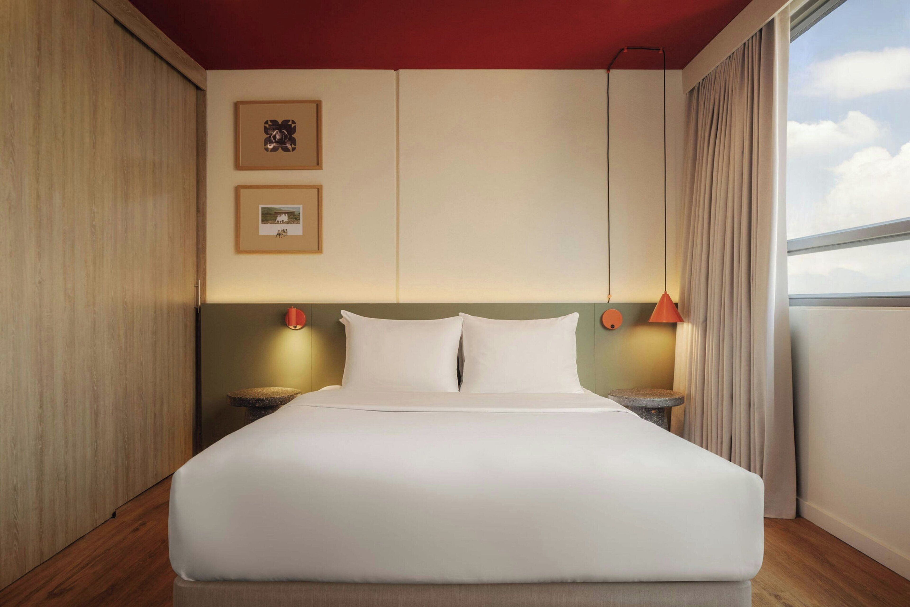 Habitación de hotel con una cama grande, dos mesitas de noche, un cabecero, una ventana con cortinas y cuadros enmarcados en la pared.