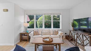 A living room with a sofa, coffee table, and TV. There are windows with a view of greenery and an American flag. - Walk to Newport, 1st & 2nd Beaches, & Cliff Walk from this Newly Redone Home (Middletown)