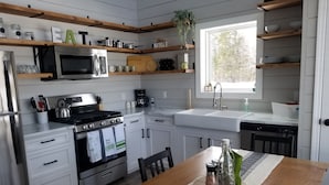 A modern kitchen with white cabinets, stainless steel appliances, wooden shelves, and a dining table with a placemat. - #5 Scenic Aspy Mountain Cottage, Margaree River access, near Cabot Trail (Margaree Valley)