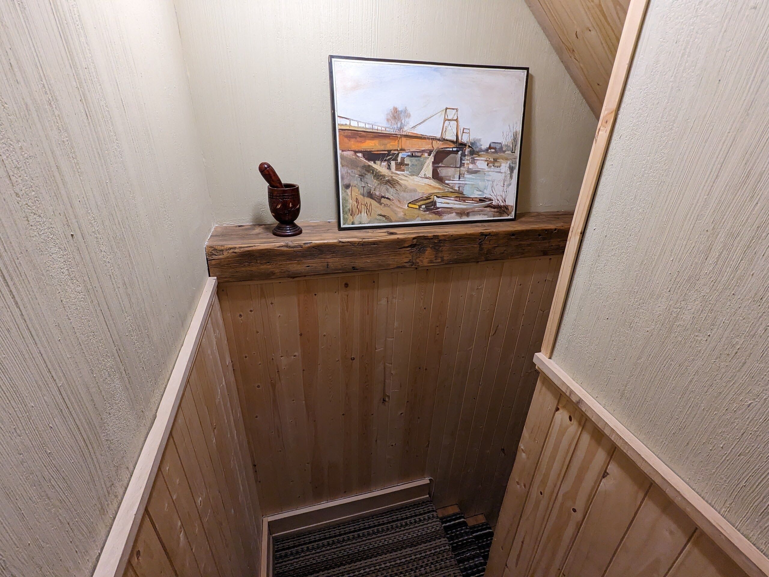 Un llarg escaler de fusta amb barana de fusta i reixeta metàl·lica a la base. Damunt de l'escaler, penjant a la paret, hi ha un quadre emmarcat d'un pont.