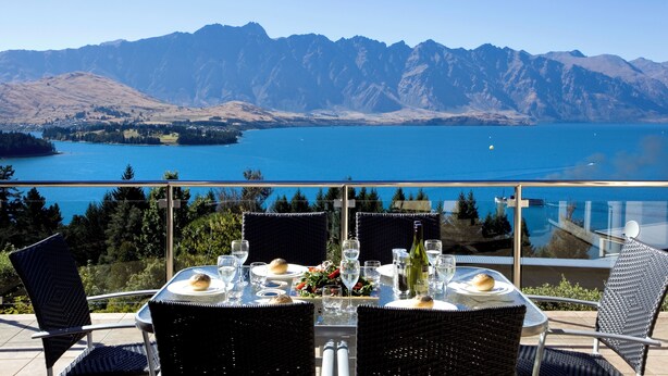 A dining table set with plates, glasses, and a bottle, overlooking a lake and mountains. - Aspen House (Queenstown)