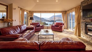 A spacious living room with leather sofas, a coffee table, and a fireplace. Large windows offer a scenic mountain and lake view. - Aspen House (Queenstown)