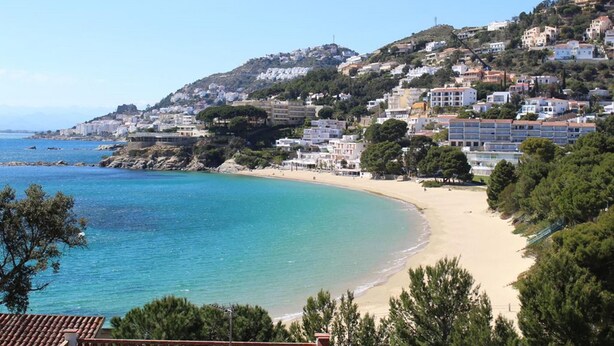 Una località costiera con spiaggia sabbiosa, edifici su una collina e mare cristallino.