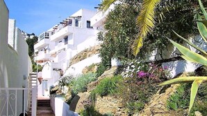 Un sentiero stretto che conduce a un edificio bianco con balconi, immerso nel verde e tra rocce.