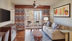 A hotel room with a flat-screen TV, a sofa, a coffee table, a dining area with chairs, a balcony with a view of tall buildings, and a large painting on the wall. - Grand Waikikian 1BR Partial Oceanview Villa (Kapolei)