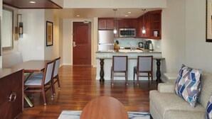 A modern living room with a sofa, coffee table, and dining area with chairs. A kitchen with wooden cabinets and a microwave is visible in the background. - Grand Waikikian 1BR Partial Oceanview Villa (Kapolei)