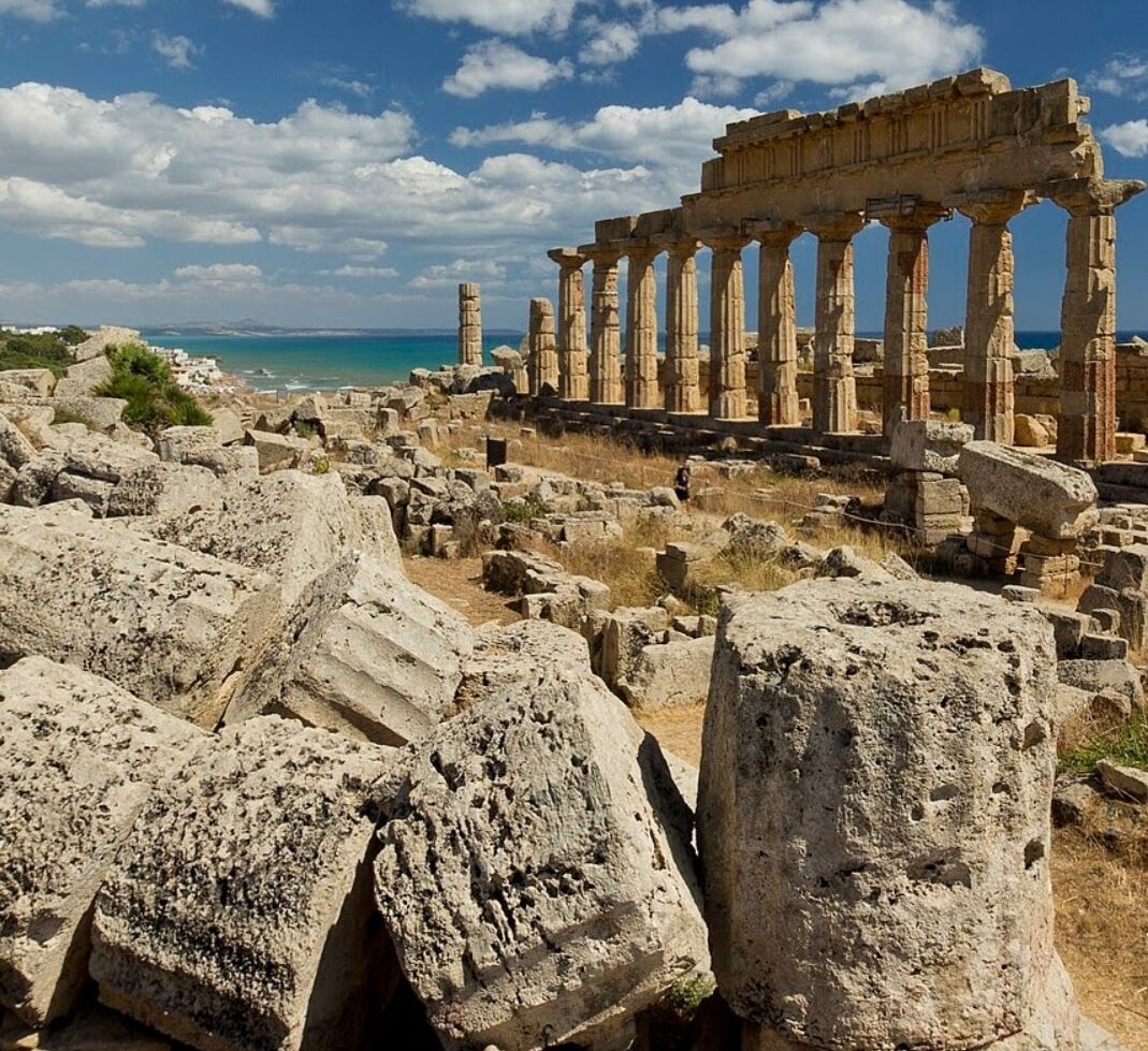 Ruinas antiguas con columnas y bloques de piedra, con vista a una masa de agua.