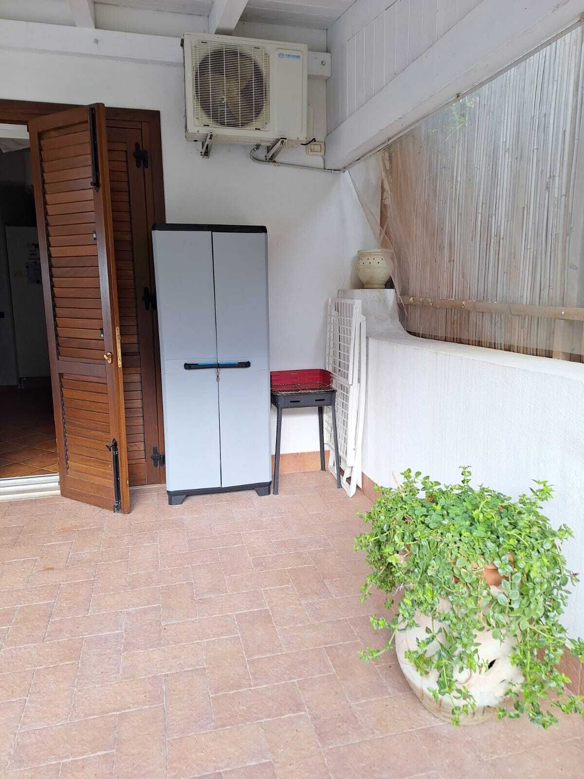Une pièce de taille modeste, avec un réfrigérateur blanc, une porte en bois, une petite table surmontée d’un coussin rouge, une plante en pot et un appareil de climatisation blanc.