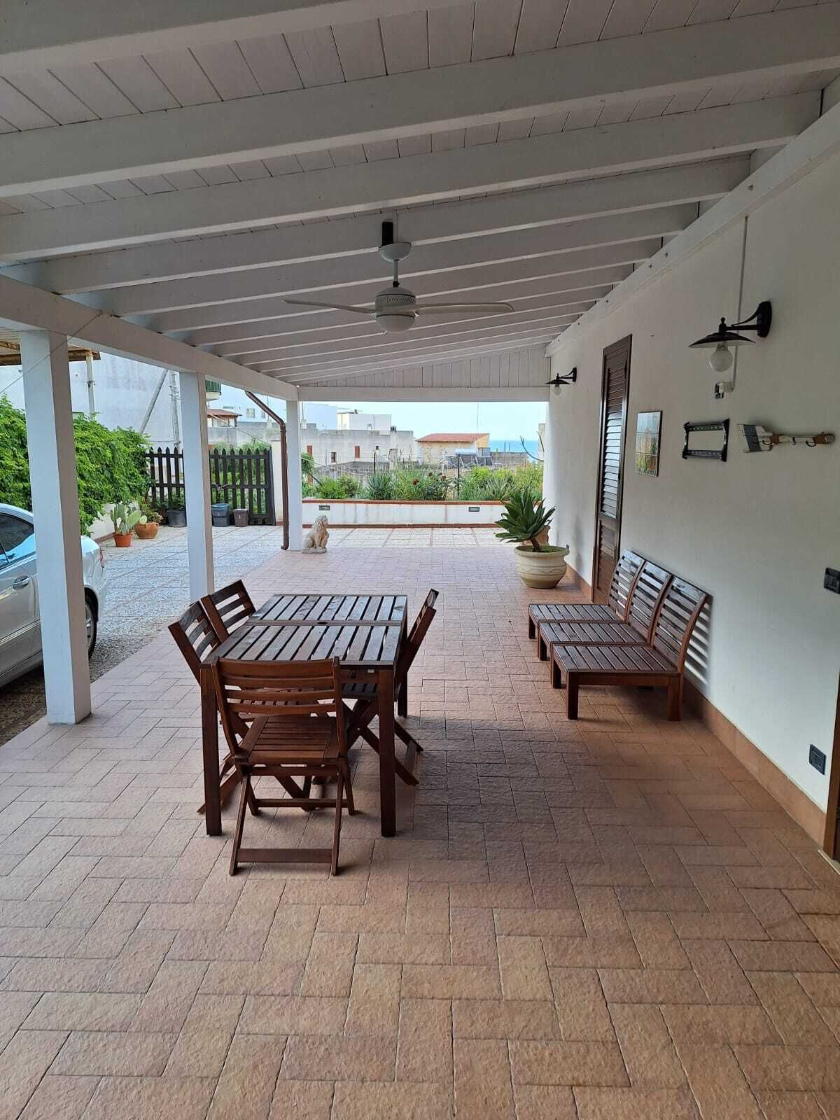 A covered patio area with a wooden table and benches, potted plants, and a view of the surrounding area.