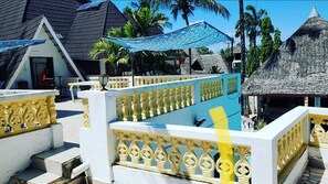 Guest rooms offer a balcony with pool views, a shaded area beneath a canopy, and a tropical atmosphere.