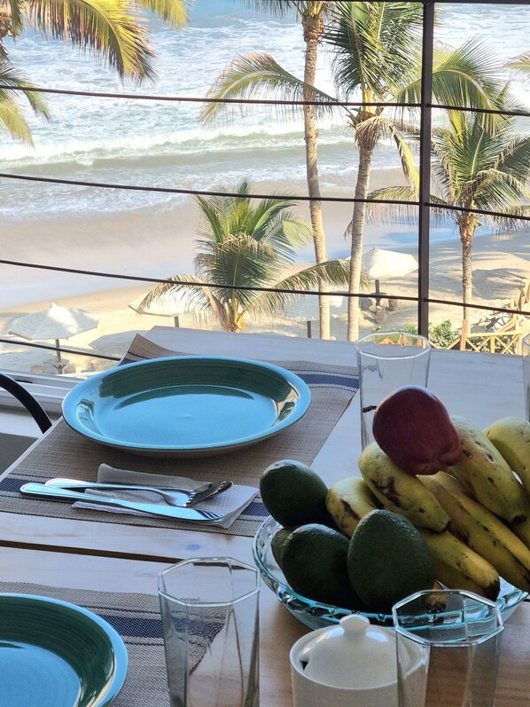 Mesa para dos con platos, cubiertos y un cuenco de fruta, con vista a una playa con palmeras y olas del océano.