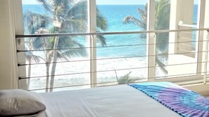 A bedroom with a large window overlooking a beach and palm trees.