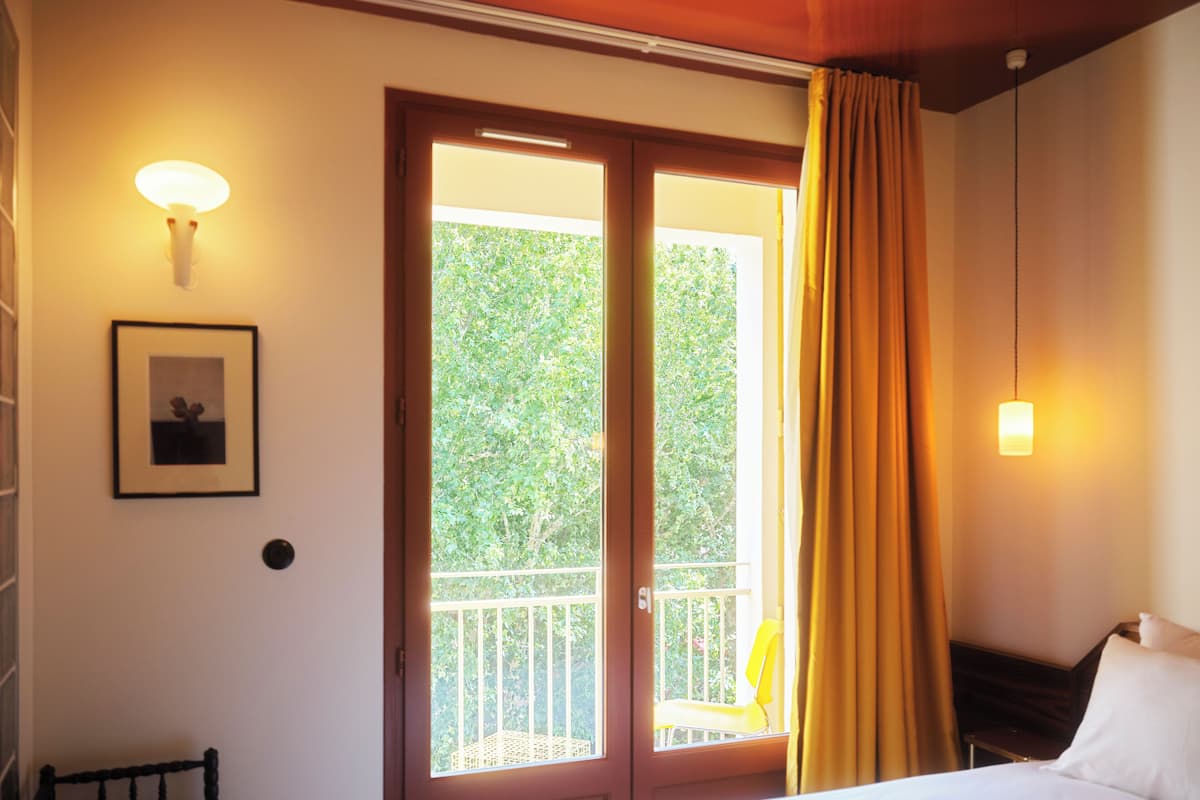 A hotel room with a bed, a chair, a picture on the wall, and a balcony with curtains.