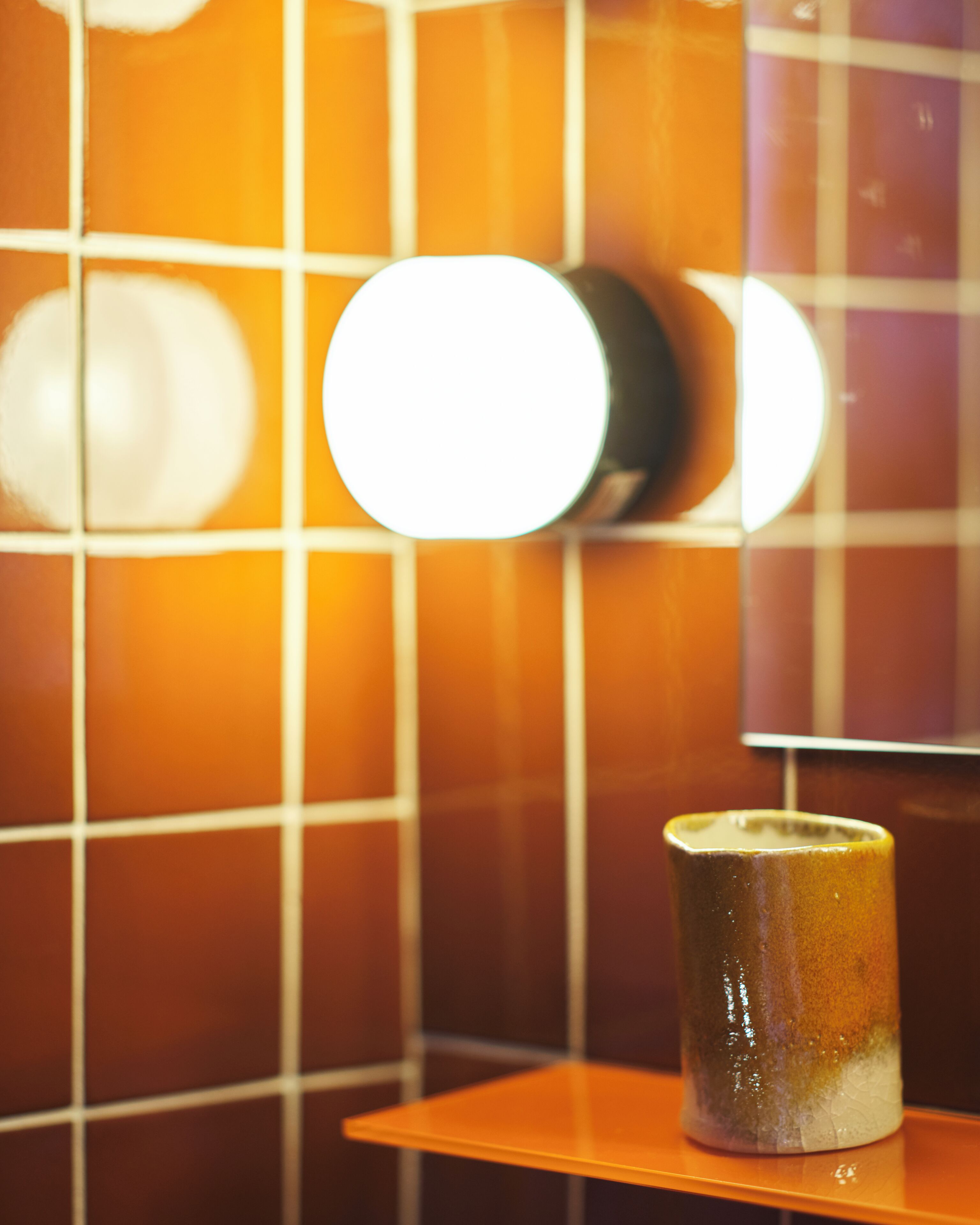 Un baño con pared de azulejos, un espejo redondo y una taza de cerámica en un estante.