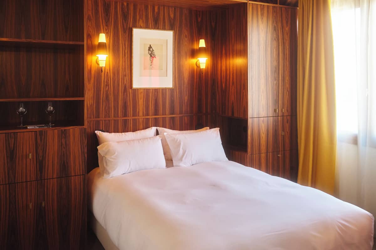 A hotel room with a wooden panel wall, a bed with white linens, two bedside lamps, and a framed picture on the wall.