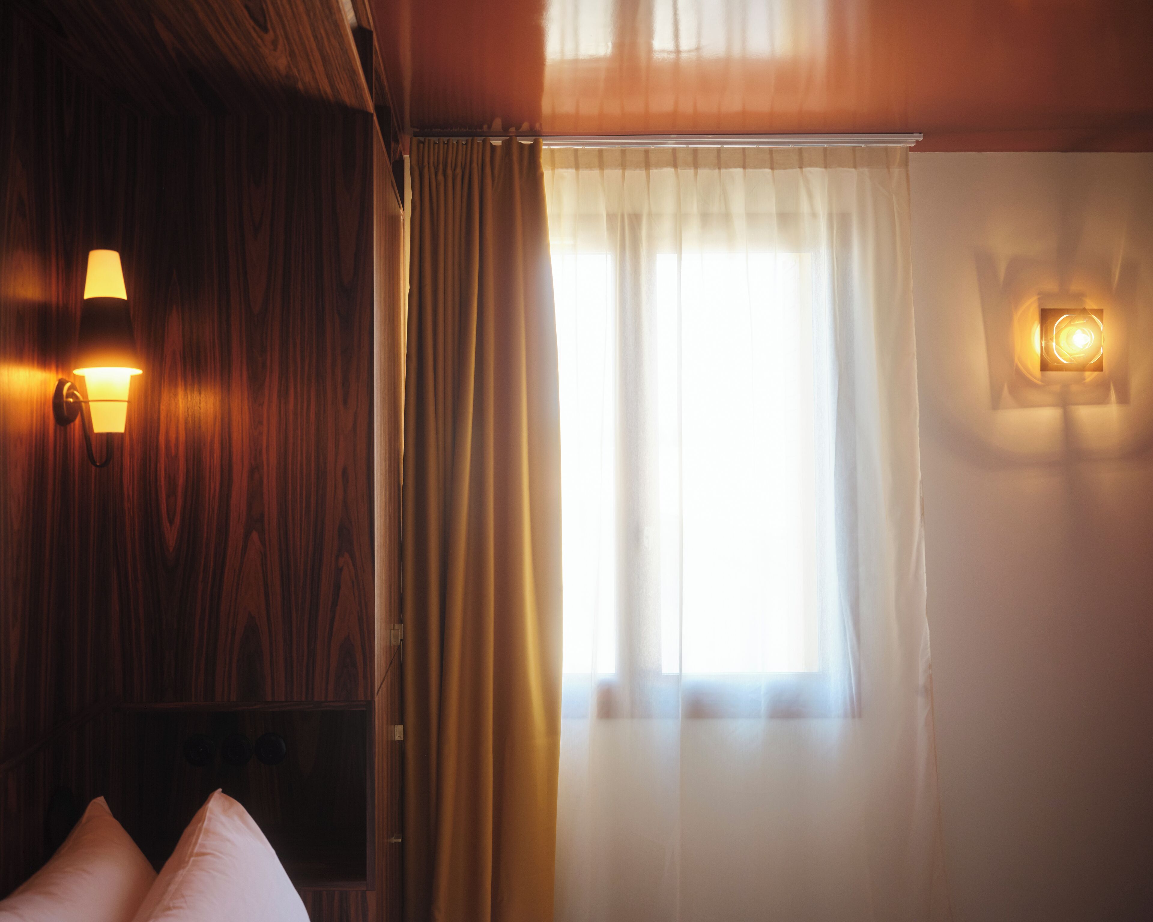 Una habitación con paneles de madera, una cama con almohadas, una ventana con cortinas e iluminación montada en la pared.
