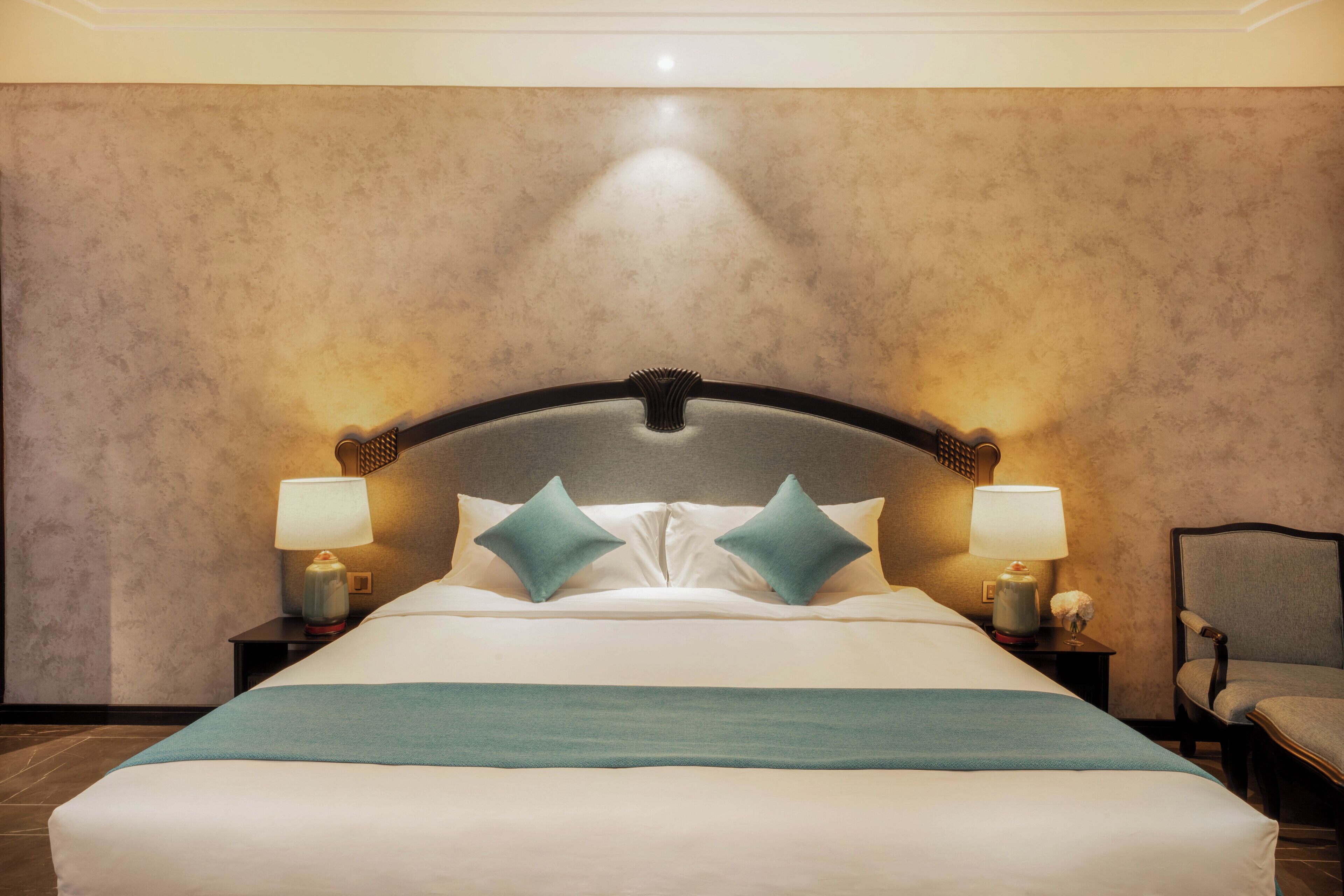 A neatly made bed with a headboard, two bedside lamps, and a chair.