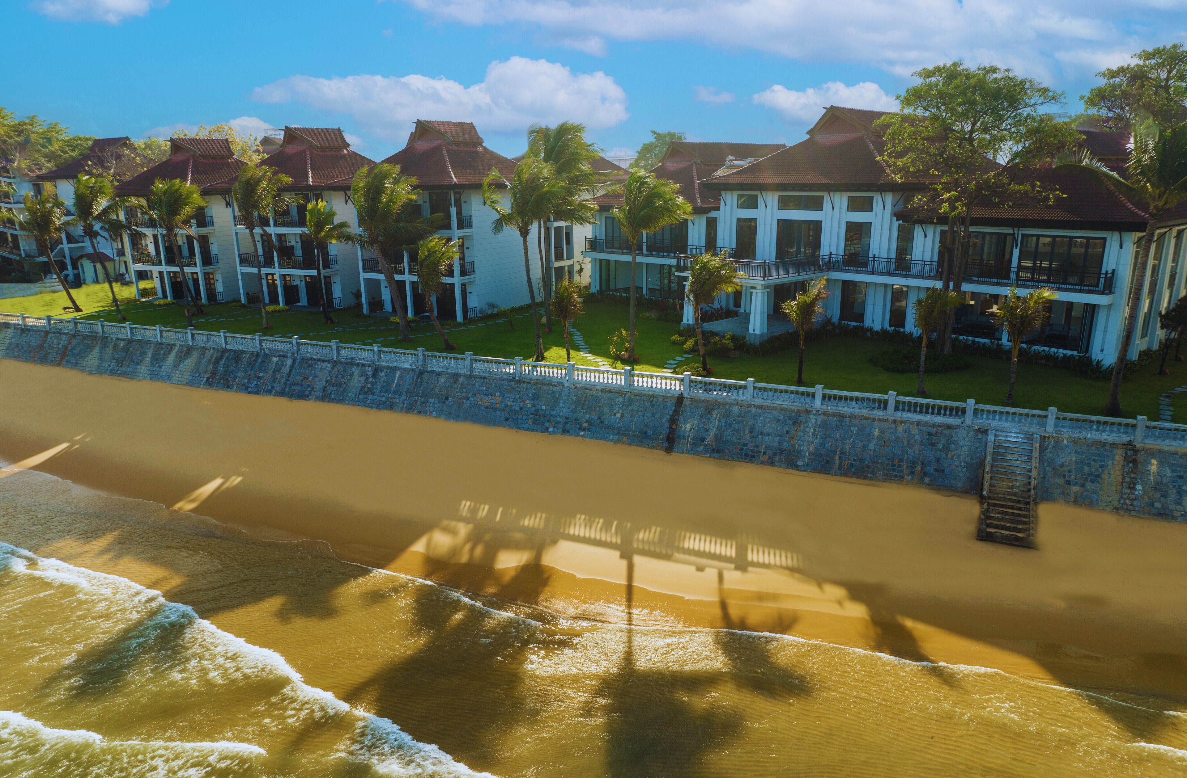 Un hôtel situé en bord de mer, composé de plusieurs bâtiments, doté d’un mur en pierre et bordé de palmiers.