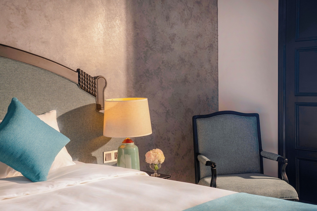 A hotel room with a bed, a chair, a lamp, and a vase with flowers.