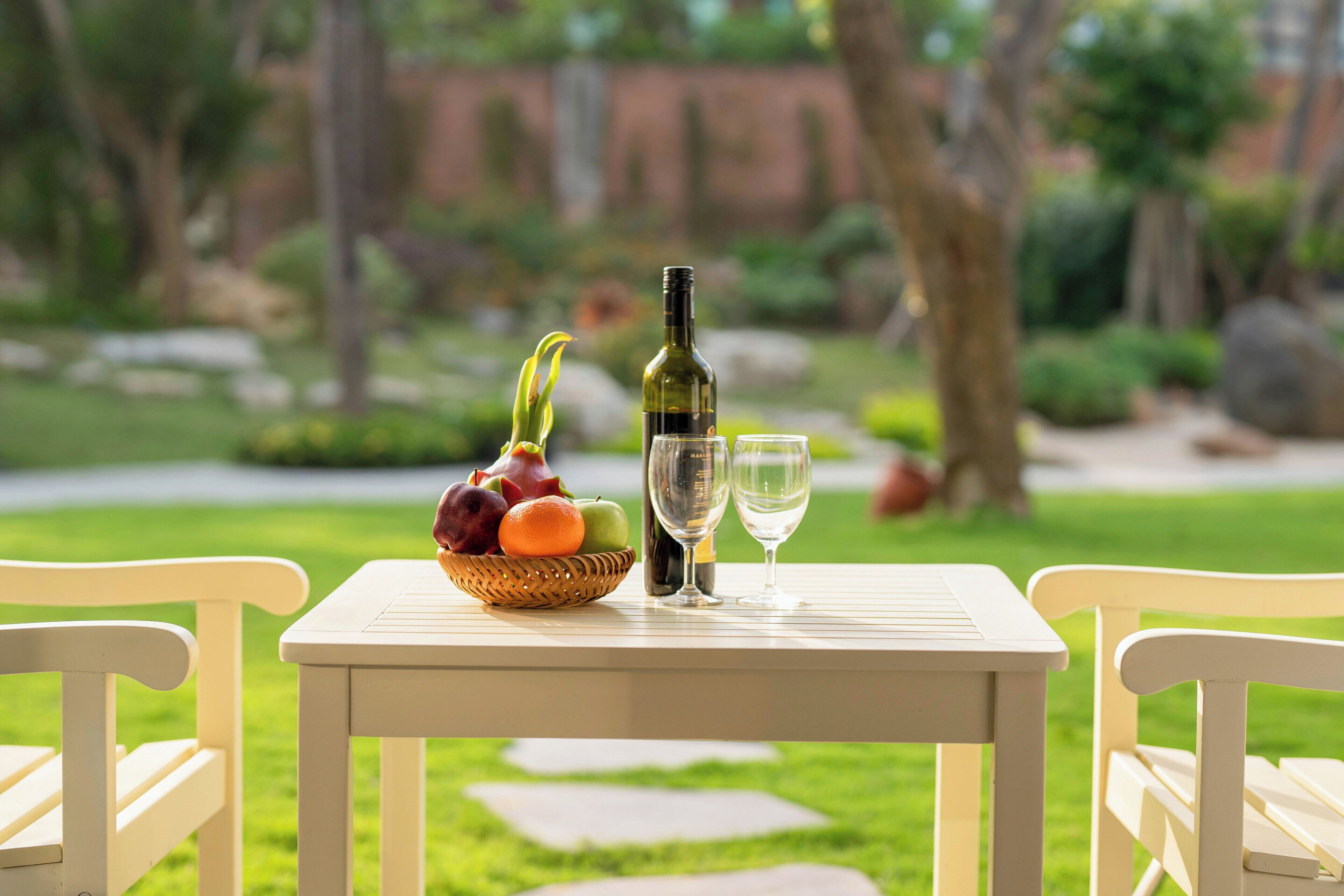 A table with a bottle, two glasses, and a fruit basket in a garden setting.
