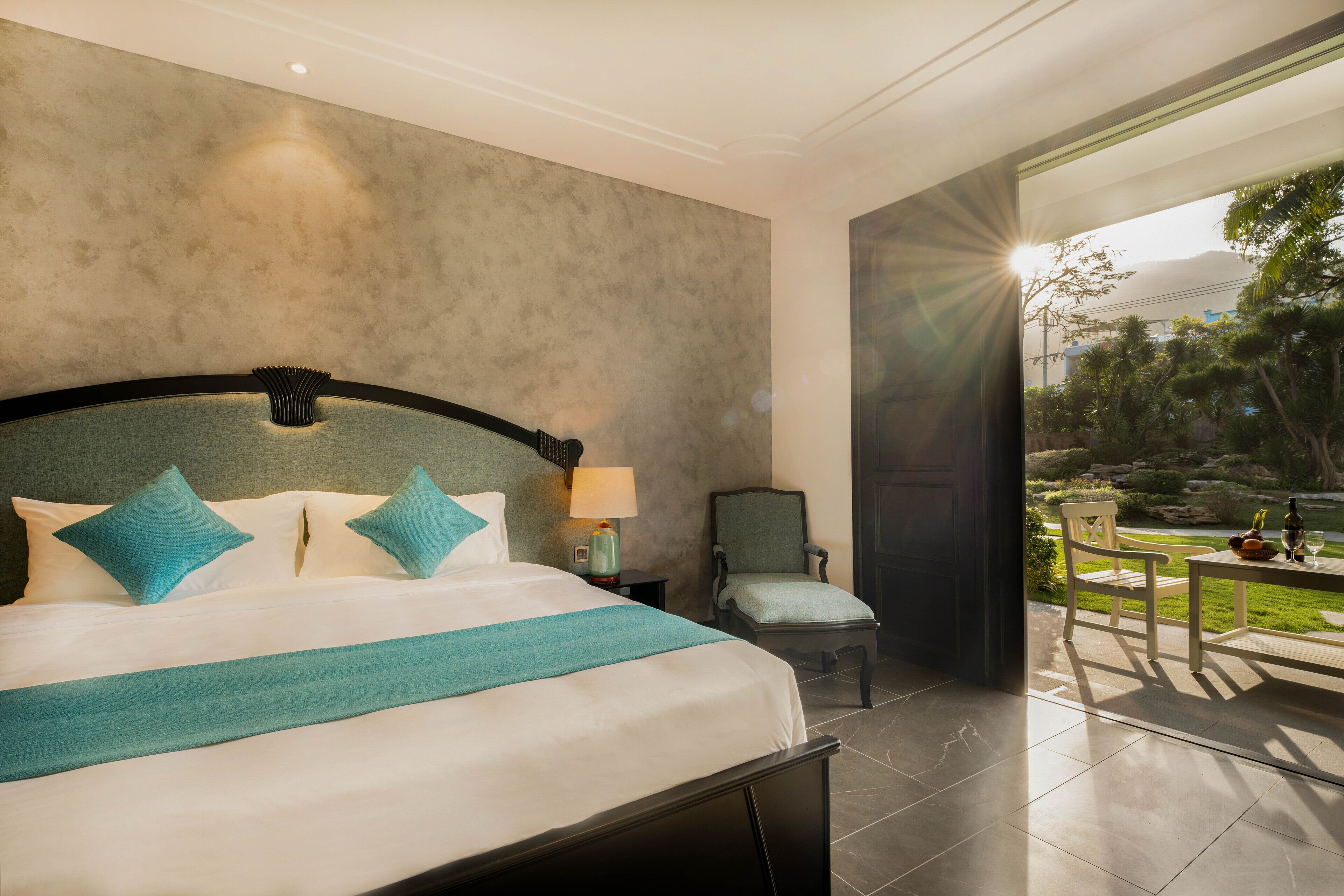 A modern hotel room with a large bed, a chair, a table with a bottle and glasses, and a view of the outdoors.