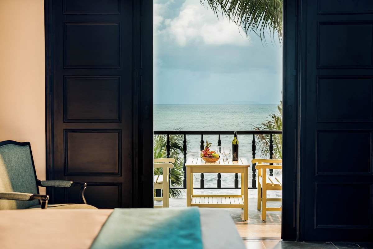 A room with a bed, a chair, a table with a bottle and fruit, and a view of the sea through an open door.