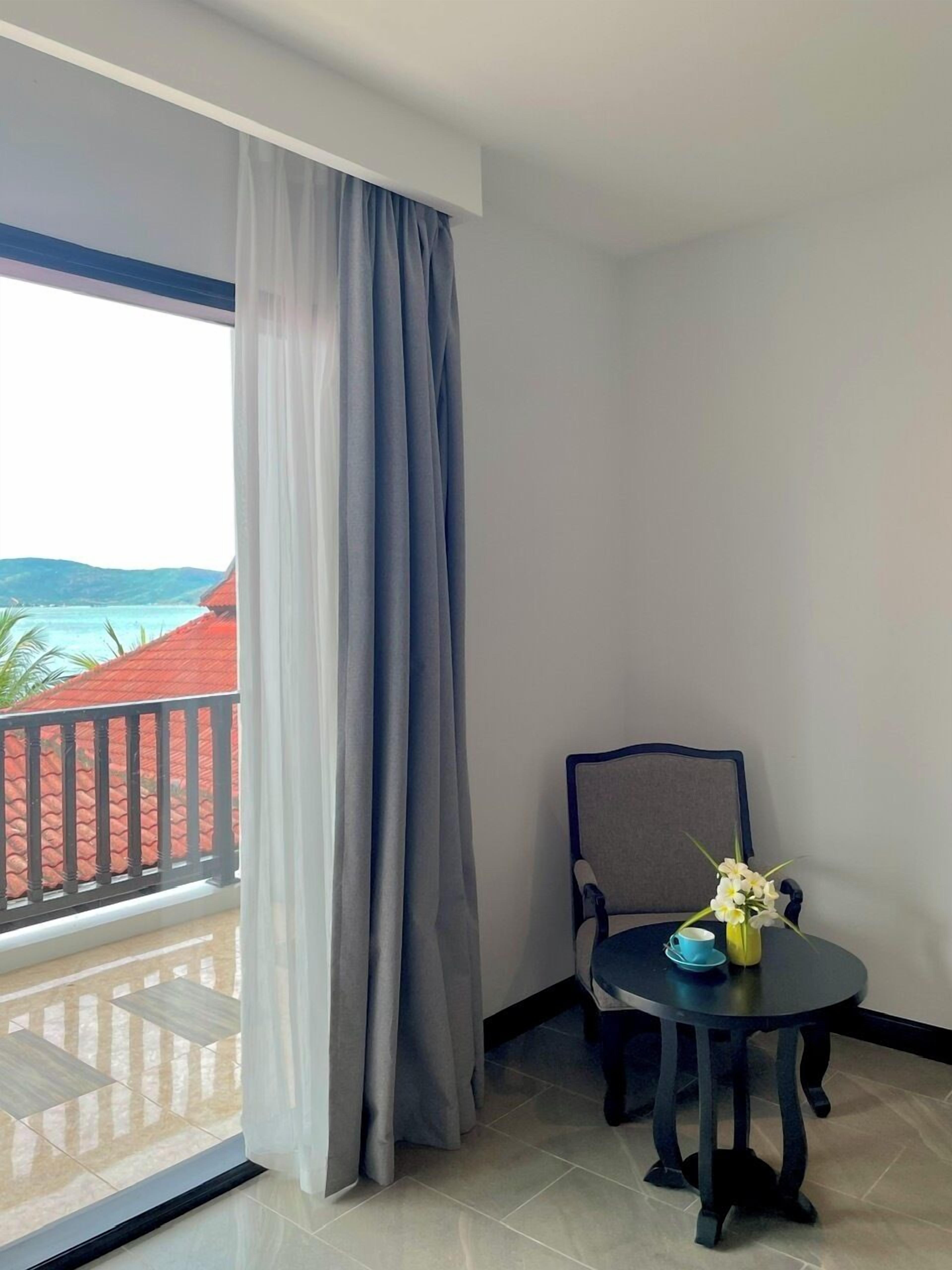 A balcony with a view of a red-roofed building and a body of water. There is a chair and a small table with a vase of flowers and a cup on it.