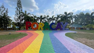 A colorful pathway leading to a sign with the word "PORACAY" in large letters, surrounded by tropical trees and a clear sky.