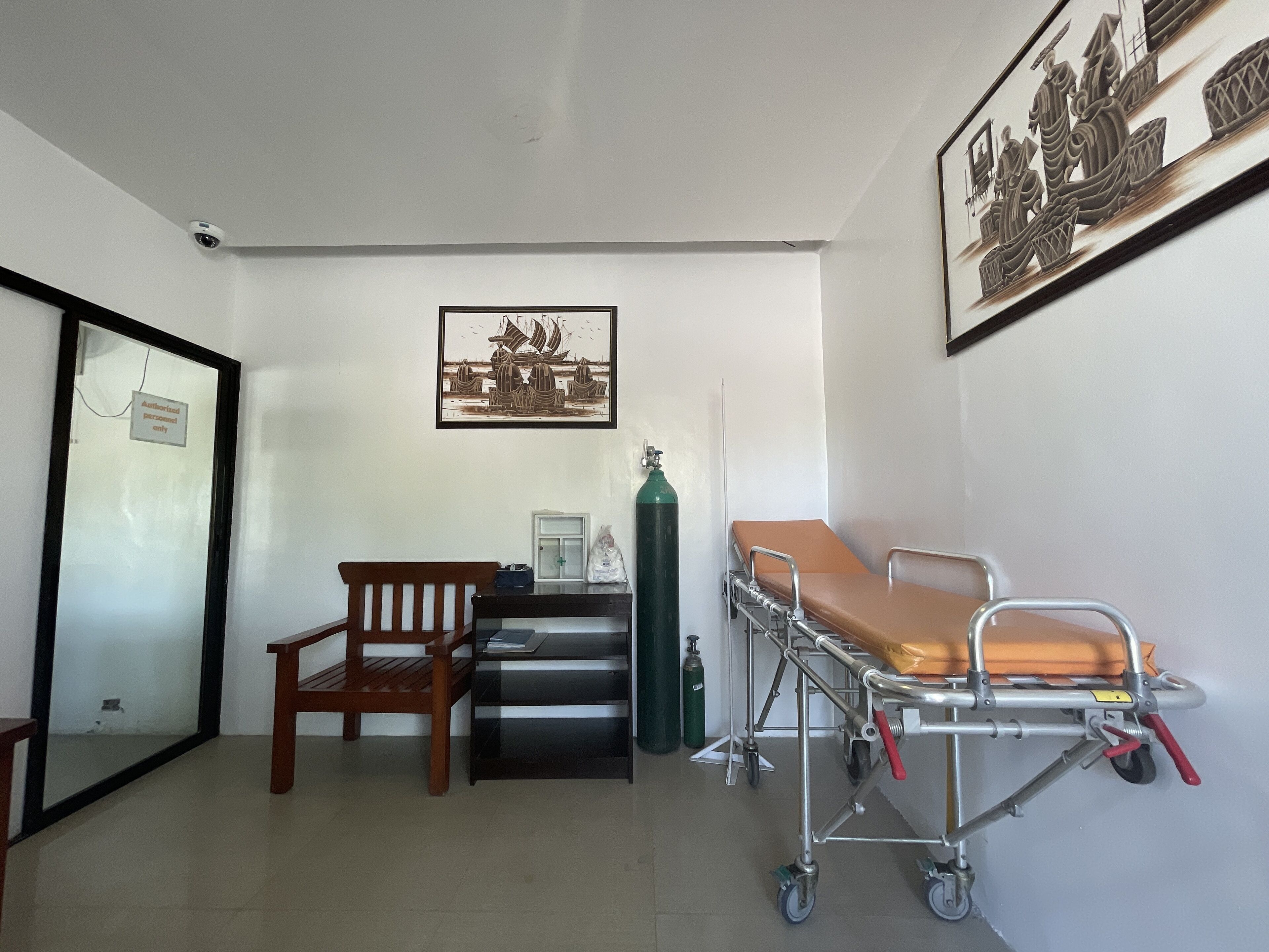 Une salle d’examen médical équipée d’un lit, d’une chaise, d’un bureau, d’une bouteille d’oxygène fixée au mur et d’un tableau représentant un navire.
