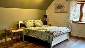 A bedroom with a sloped ceiling, a bed with a green bedspread, a wooden nightstand, and a window with curtains.