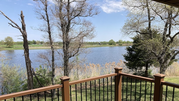 A wooden deck with a railing overlooking a lake, surrounded by trees and grassy areas.