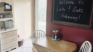 A dining area with a wooden table and white chairs, a chalkboard sign, and a kitchen area with a white cabinet and shelves.