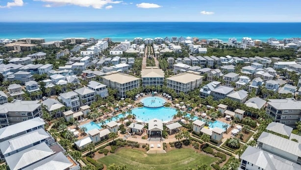 An aerial view of a resort with multiple buildings, a central pool area, and a clear view of the ocean.
